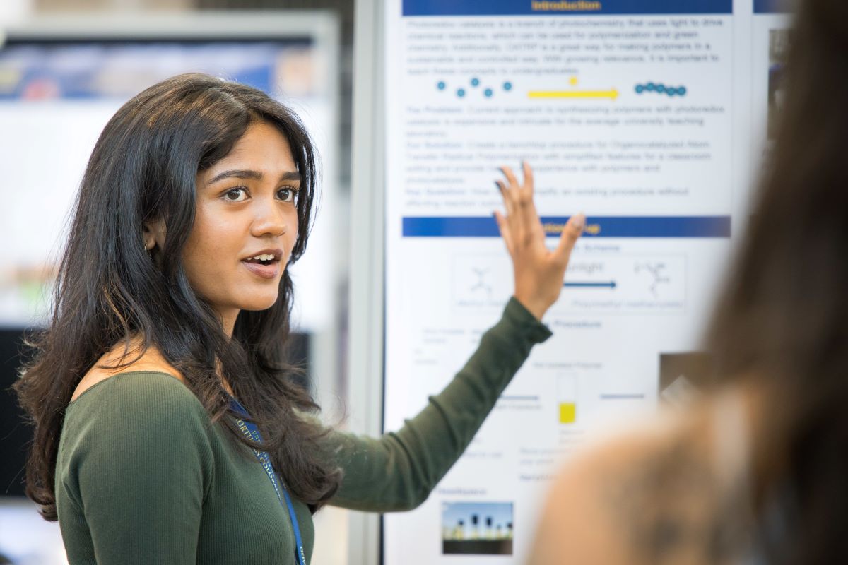 a student with dark hair and a green shirt stands in front of a research poster