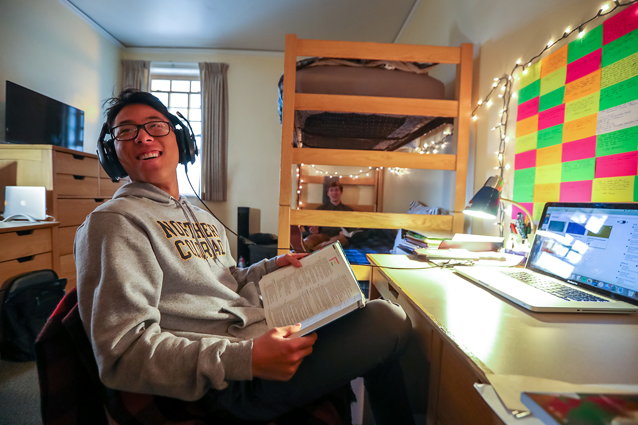 Student smiles while sitting at their desk holding a textbook in their new dorm room.