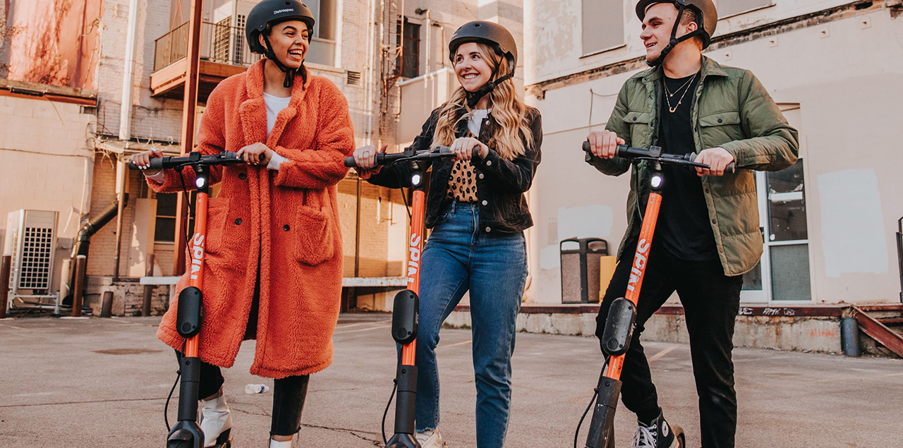 Three college-age people standing outside beside e-scooters.