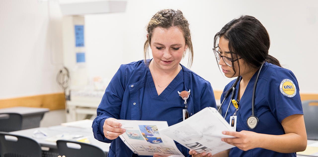 Two nursing students in blue scrubs standing side-by-side reading course materials