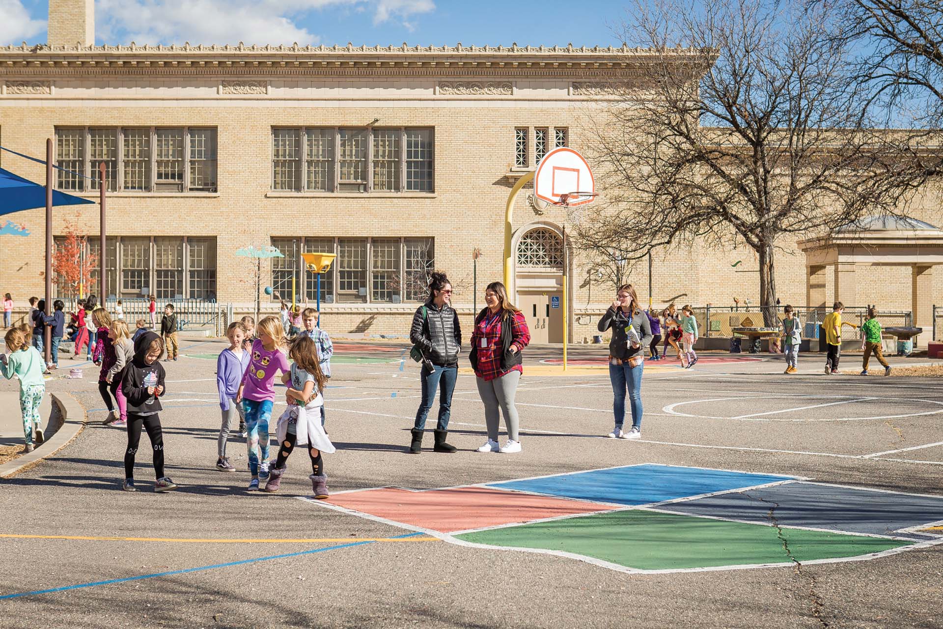 Two teachers stand on a basketball court of an elementary school during recess.
